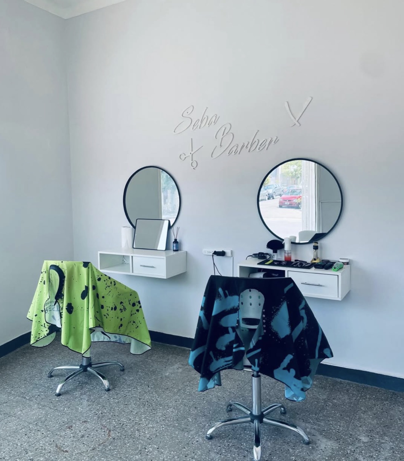 Interior de Sebas Perez Barber: sillas con capas de colores, espejos redondos y la pared con el nombre Seba Barber.